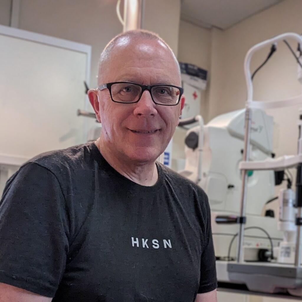 Gary Optometrist sat looking towards camera , wearing a black tshirt with HKSN typed accross. In the background are various optical instrucments